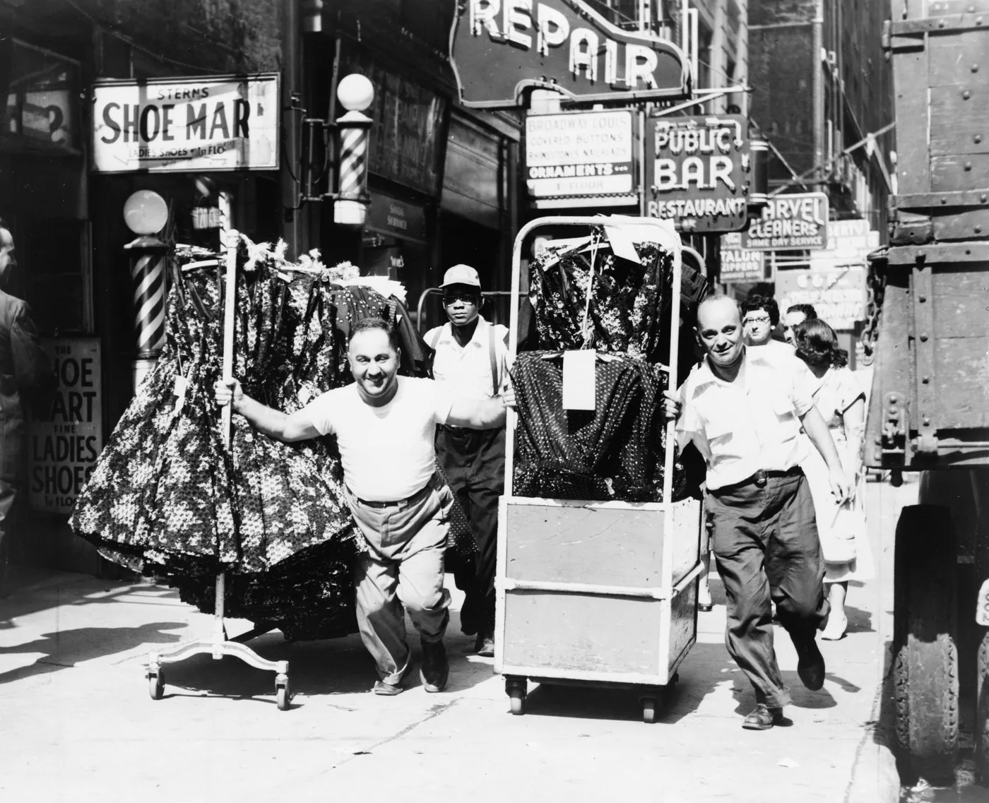 Three men rolling two racks of dresses along a crowded sidewalk in New York’s Garment District, shop signs overhead