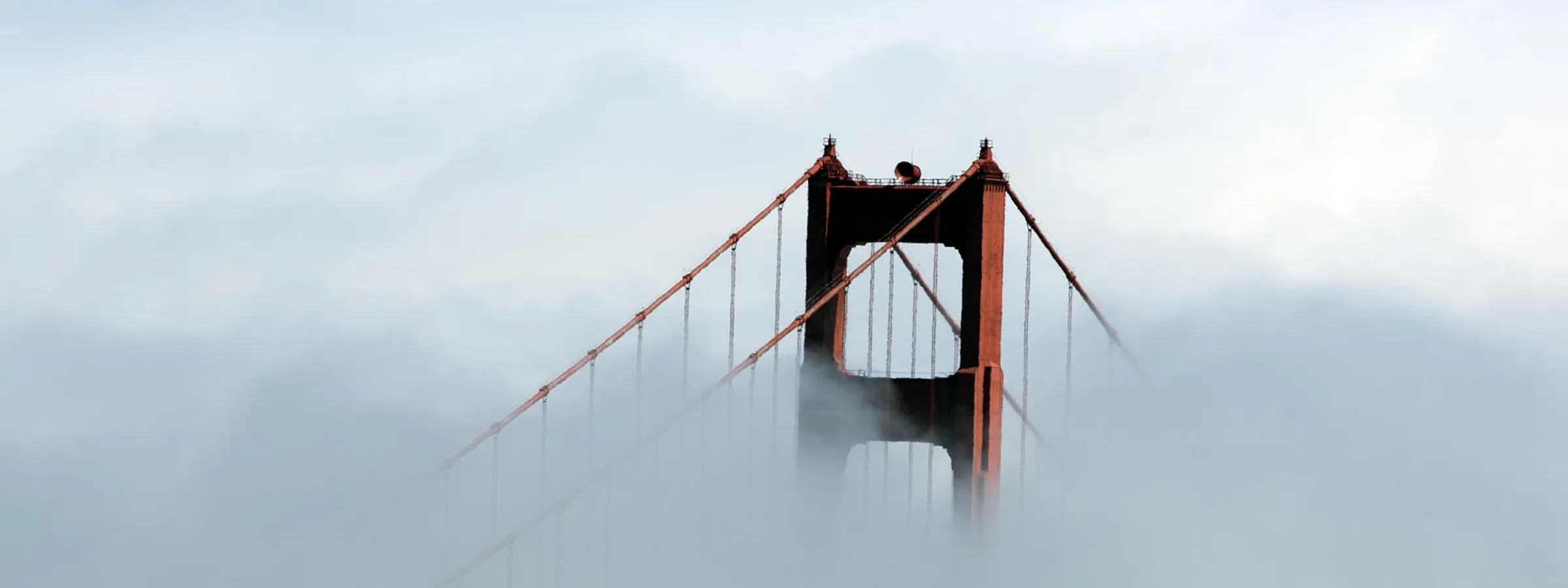 A tower of the Golden Gate Bridge rising out of a bank of fog, its red cables running down into the gray